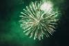 A photograph showing a green firework exploding in the night sky, surrounding by smoke