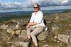 Jill is sitting on some rocks, wearing sunglasses and smiling. There are hills in the background. 