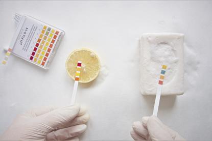 A scientist wearing disposable gloves holds indicator paper over a slice of lemon and a bar of soap