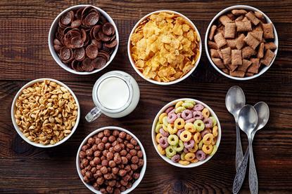 A variety of different breakfast cereals in bowls on a wooden surface, with a glass jug of milk and spoons at the side