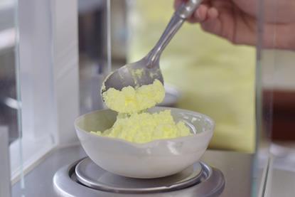 A close up of someone using a spoon to add sulfur powder to a weighing boat on top of a balance