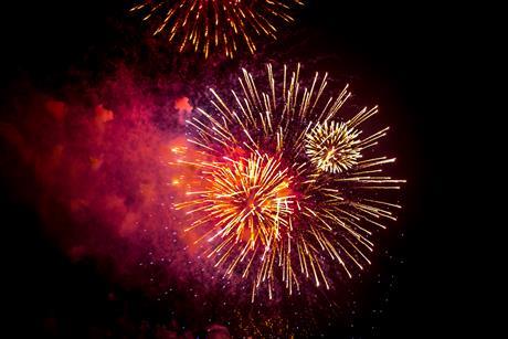 A photograph of red, orange and gold fireworks exploding against the night sky, giving off smoke that appears to have a red colour