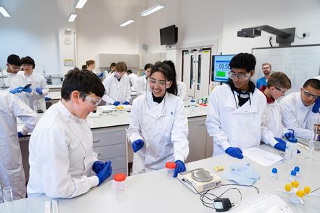 A small group of students wearing lab coats and goggles smile and discuss an experiment at a laboratory bench, while other groups work in the background